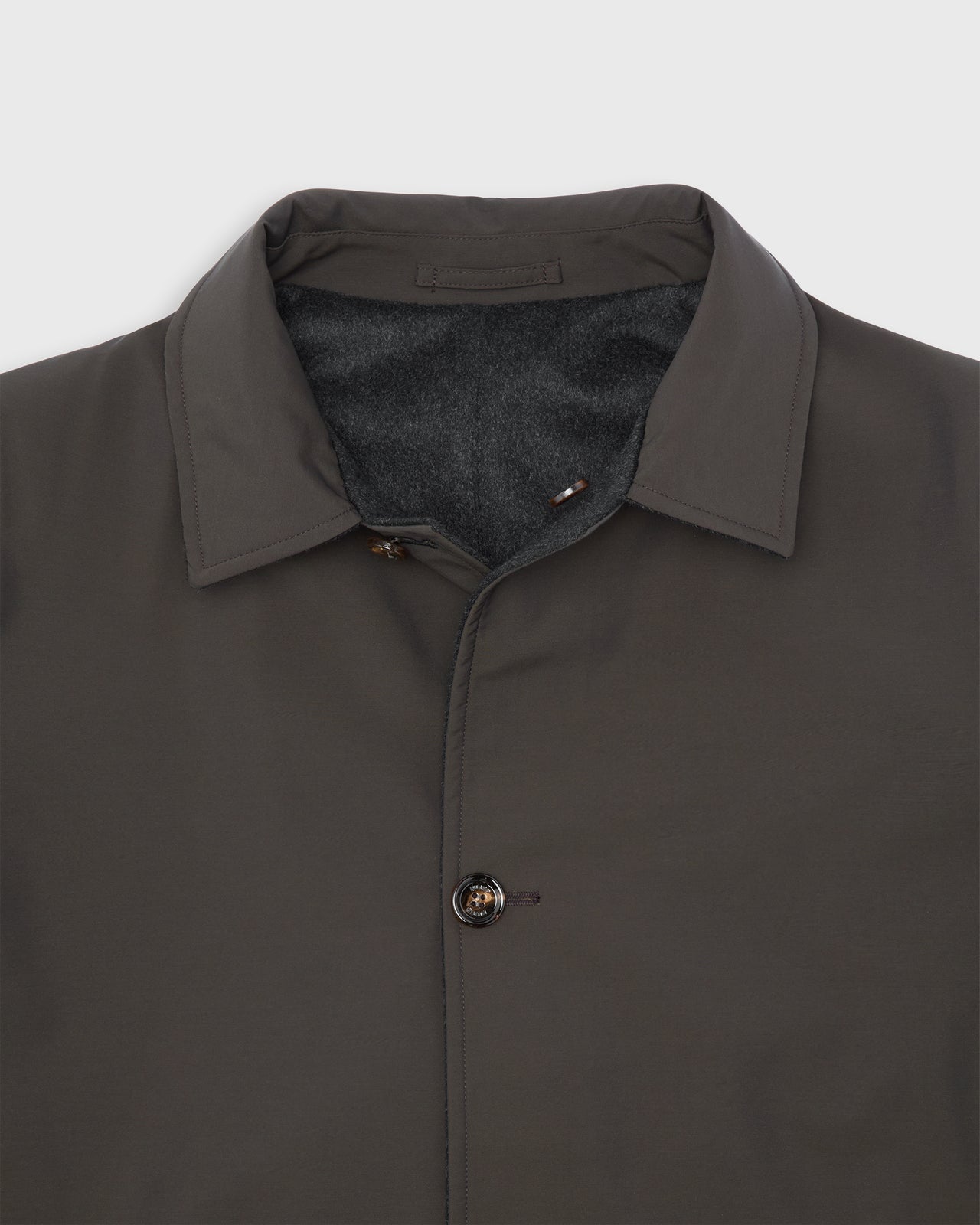 Close-up of a dark brown collared jacket with a dark gray cashmere reversible lining and a single breasted closure, shown against a light gray background.